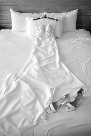 Black and white photo of a wedding dress on a hanger laid on a bed with white heels beside it.