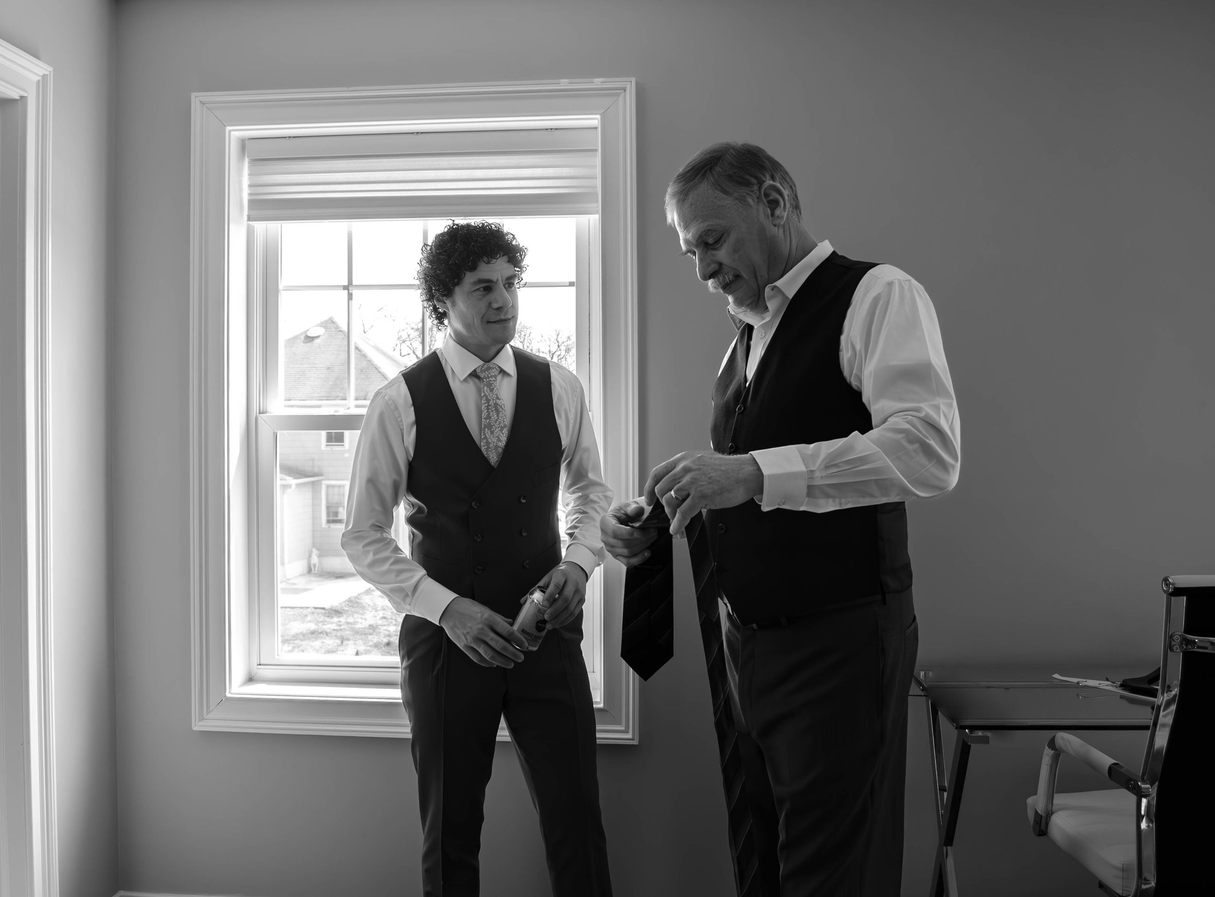 Two men in formal vests and dress shirts prepare neckties indoors in black and white photo.