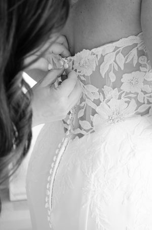 Black and white photo of hands fastening delicate lace buttons on the back of a bridal gown.