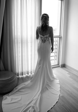 Black and white photo of a bride in a fitted wedding gown with lace back, standing before a sheer curtained window.