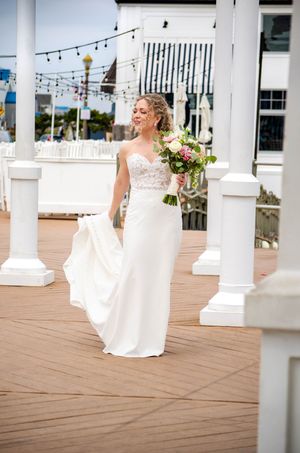 Bride in a strapless lace wedding gown holding a floral bouquet walks on a wooden deck at a waterfront venue.