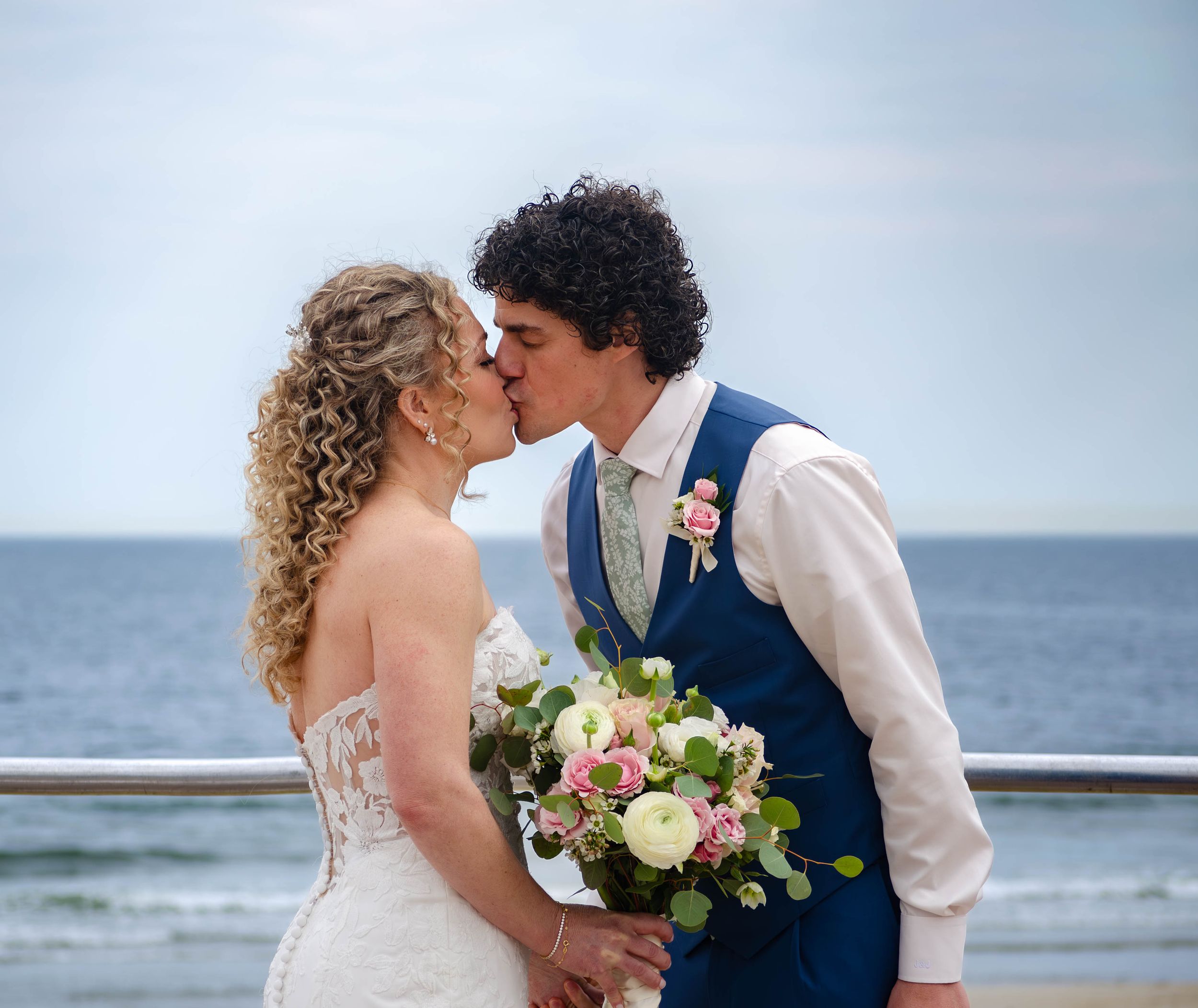 Bride and groom share a romantic kiss by the ocean, bride holding a bouquet of white and pink roses in wedding attire.