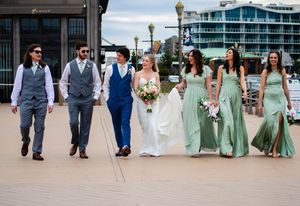 Wedding party walks outdoors, bride in white gown, groom in blue suit, bridesmaids in sage green dresses, groomsmen in gray suits.
