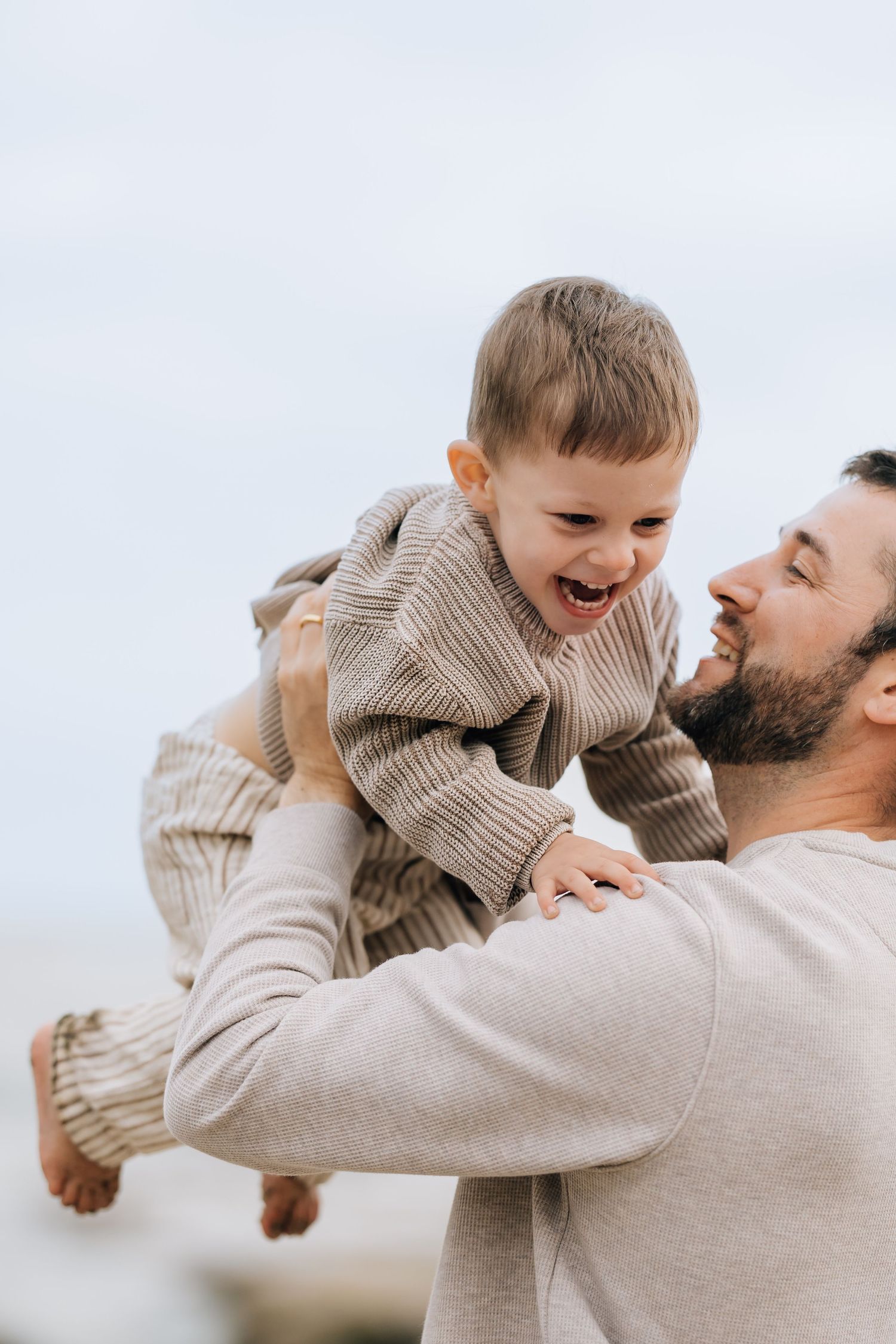 A joyful father lifts his laughing toddler in the air outdoors, both wearing matching beige knit sweaters.