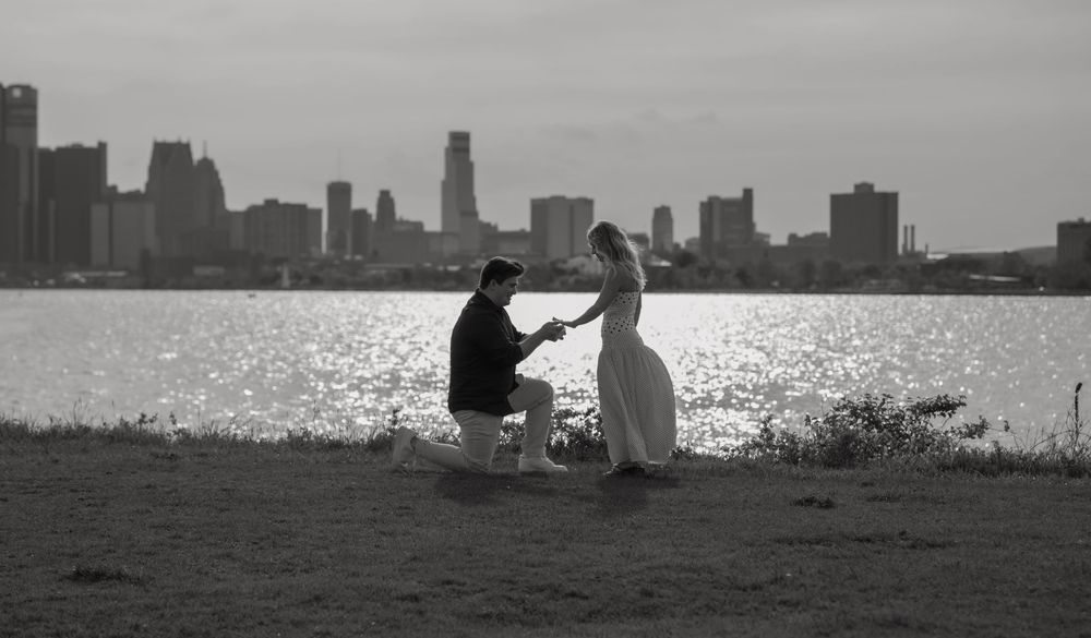 Black and white photo of a man proposing to a woman by a waterfront with a city skyline in the background.