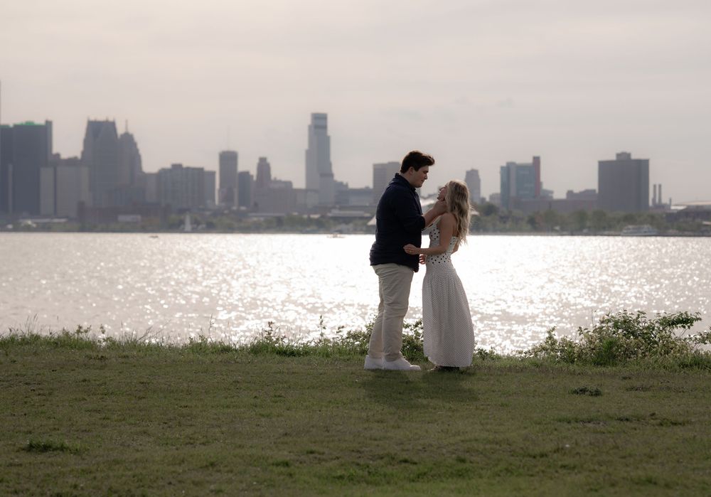 Romantic couple holding hands by a waterfront with a city skyline backdrop on a sunny day.