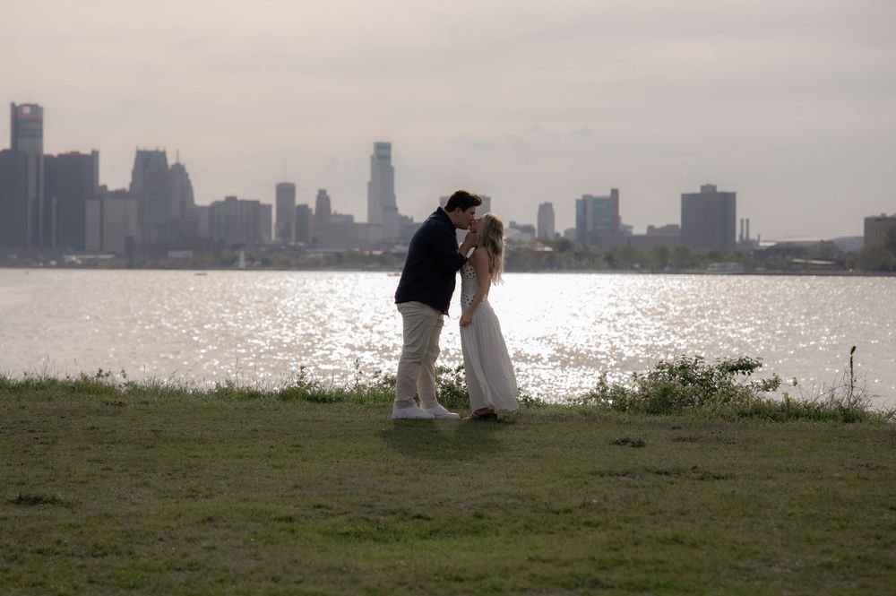 Couple sharing a romantic kiss on a grassy waterfront with a city skyline reflecting on the river behind them.