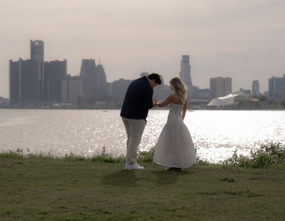 Couple in wedding attire dancing on grass by waterfront with city skyline backdrop.