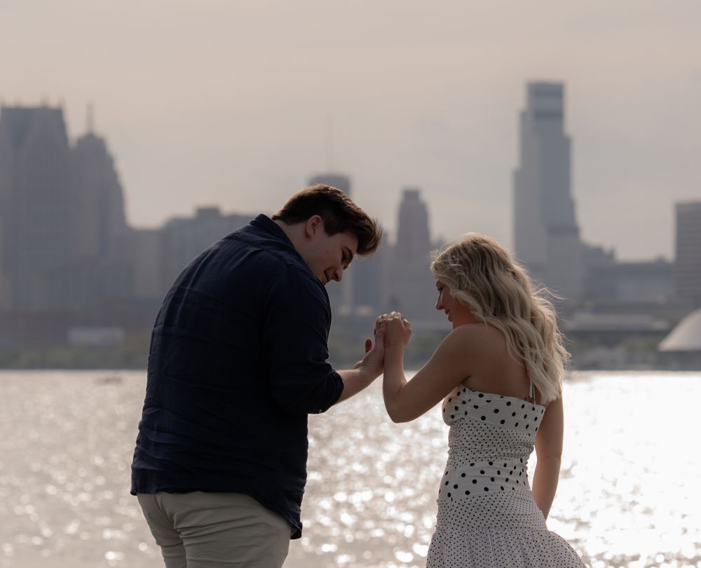 Man proposing to blonde woman in polka dot dress by waterfront with city skyline backdrop.