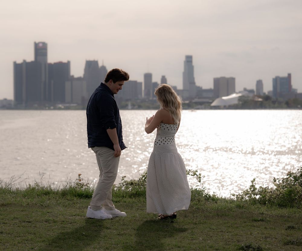 Couple standing by waterfront with city skyline backdrop during romantic outdoor engagement session.