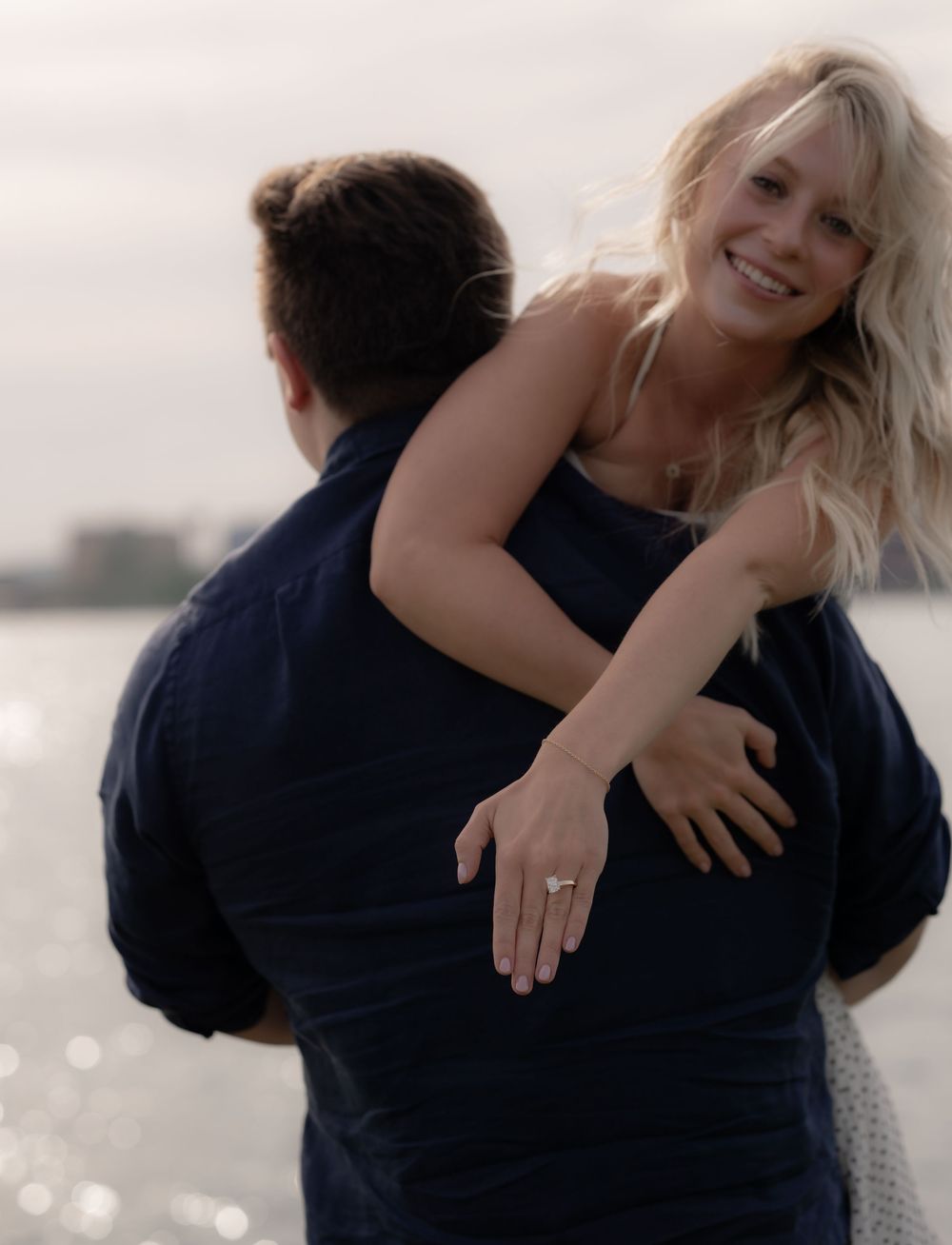 Couple celebrating engagement near water, woman showing diamond ring while being carried by man in navy shirt.