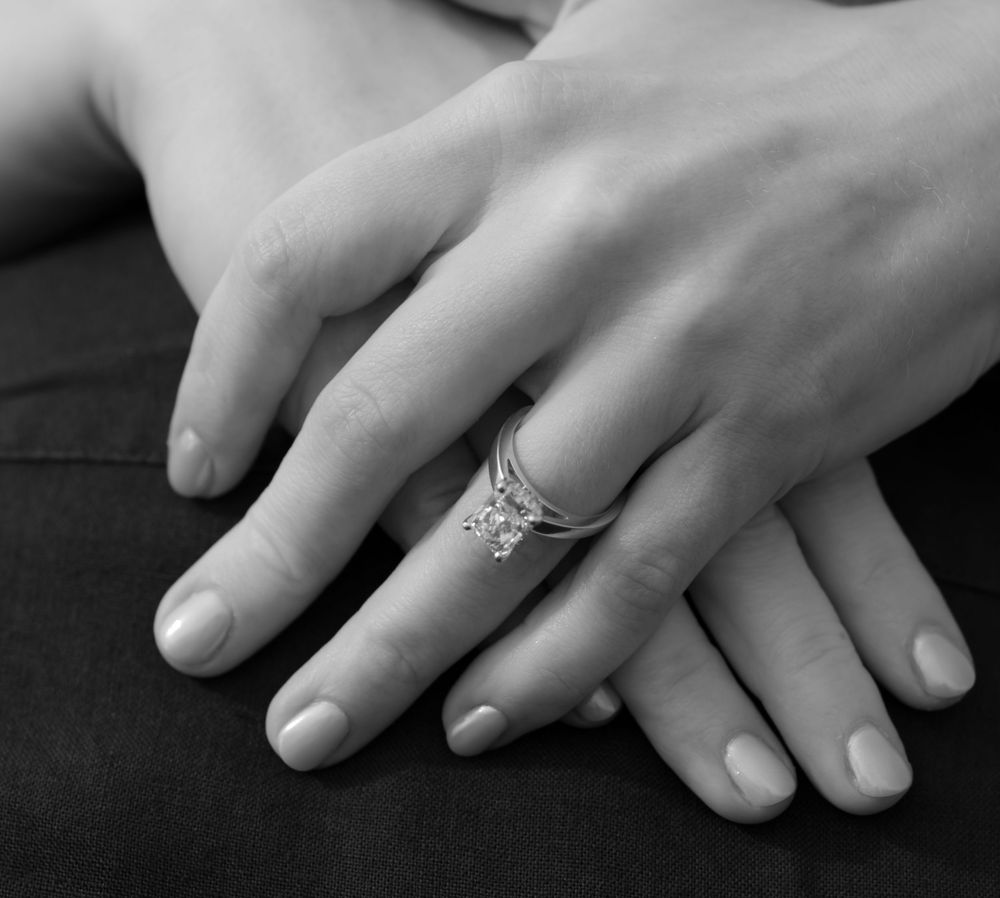 Close-up black and white photo of engagement ring with diamond on womans hand resting on partners hand.