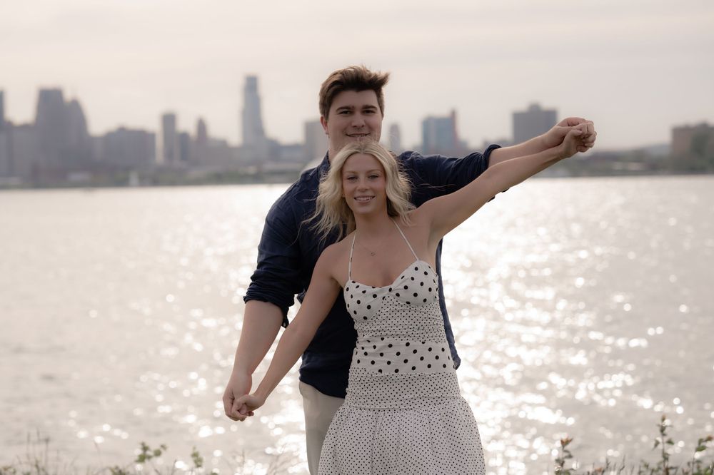 Couple posing romantically by a river with a city skyline backdrop, woman in polka dot dress spinning joyfully.