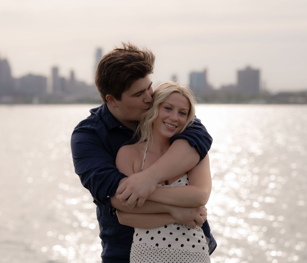 Happy couple embracing and smiling together by shimmering waterfront with city skyline in background.