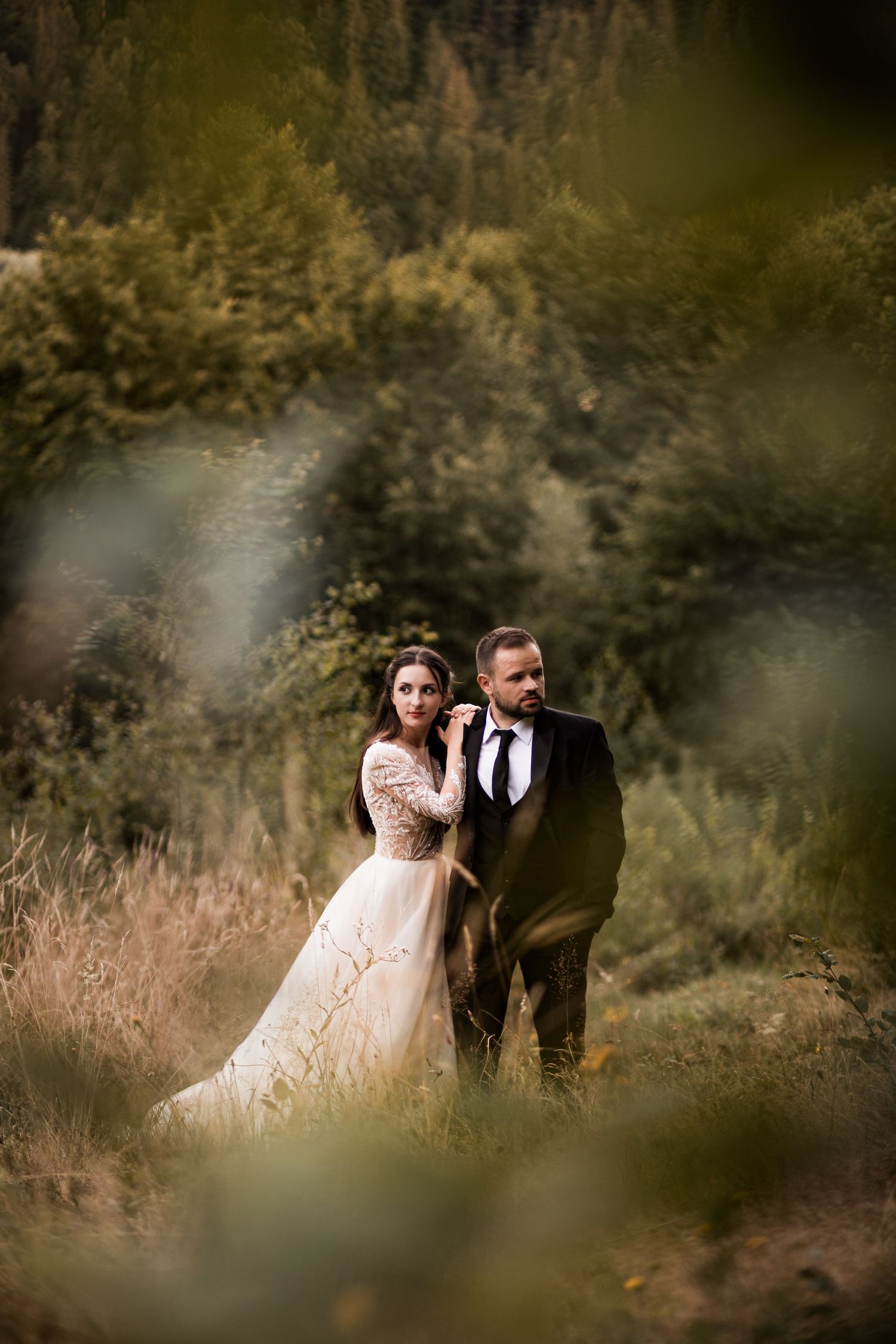 Bride in lace gown and groom in dark suit stand together in lush green forest setting during outdoor wedding photo session.