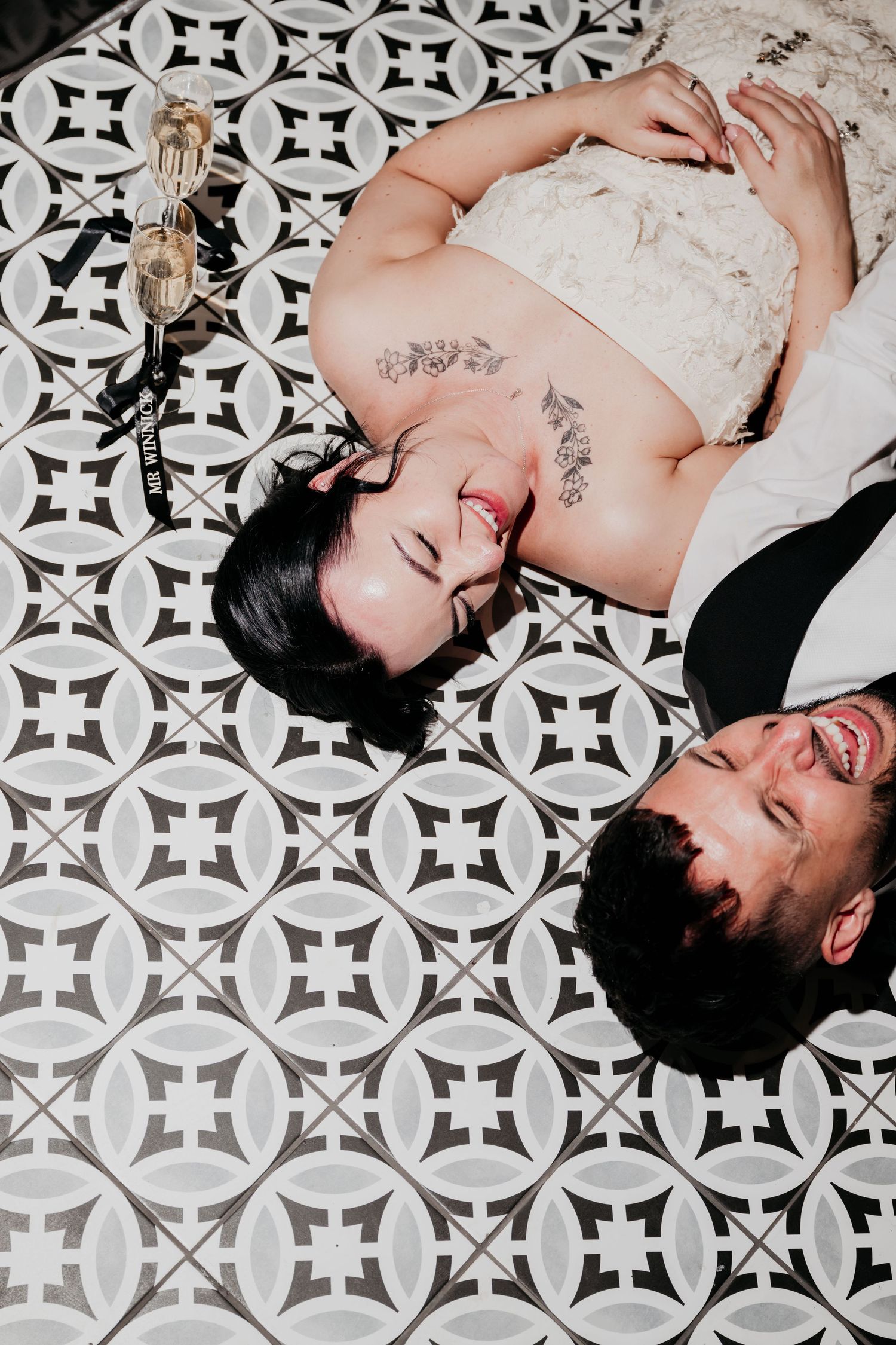 A laughing bride and groom lying on a black and white patterned tile floor with champagne glasses nearby.