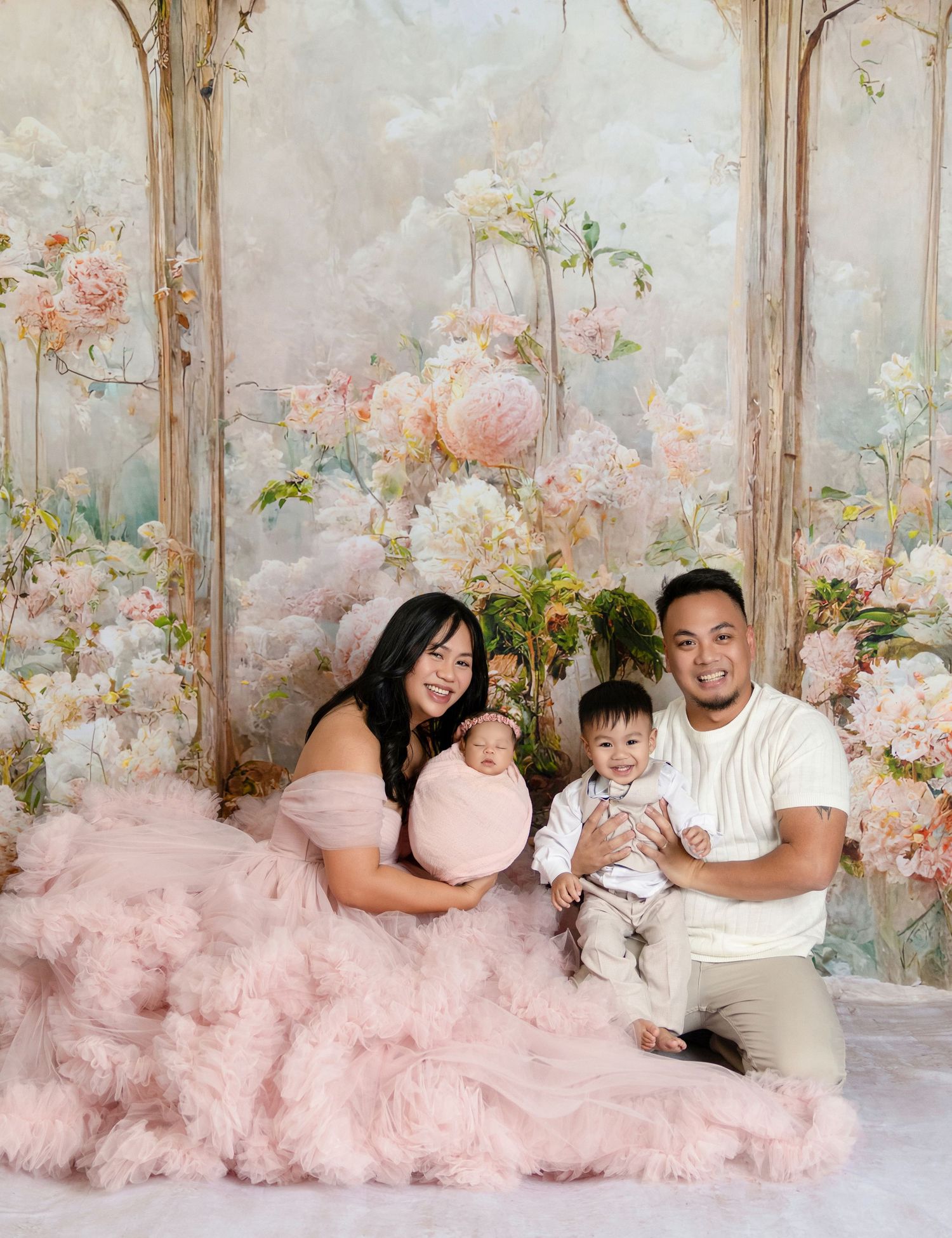 Family portrait with mom in pink tulle gown, dad, and two babies posed before a floral backdrop.
