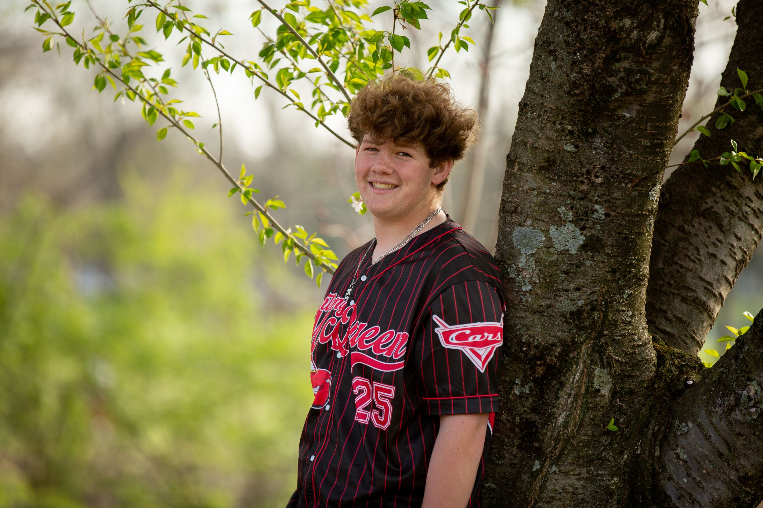 Smiling teen in black pinstripe baseball jersey number 25 posing outdoors by a tree with spring foliage.