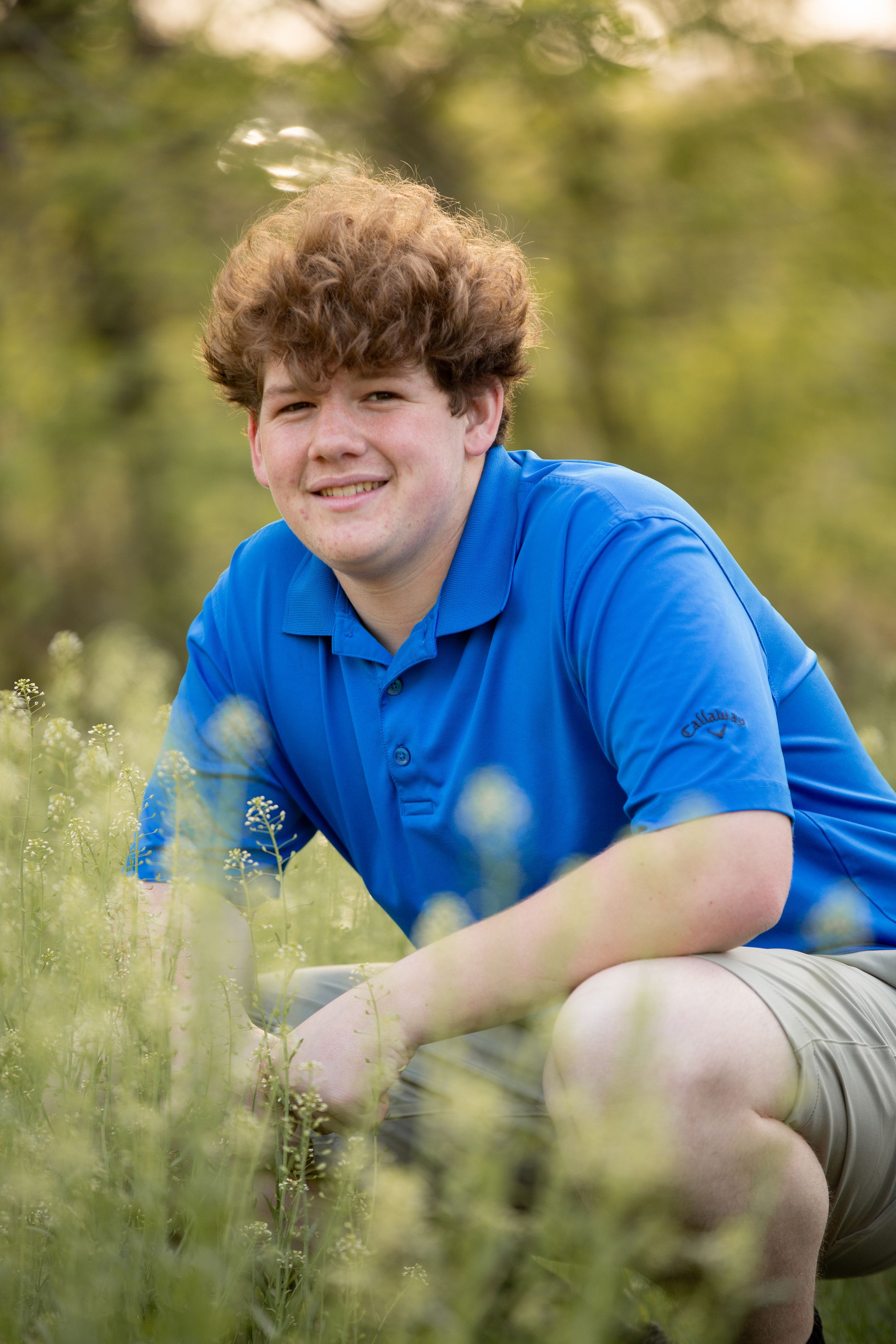 Smiling teenage boy with curly red hair crouching in a field of wildflowers, wearing a blue polo shirt.