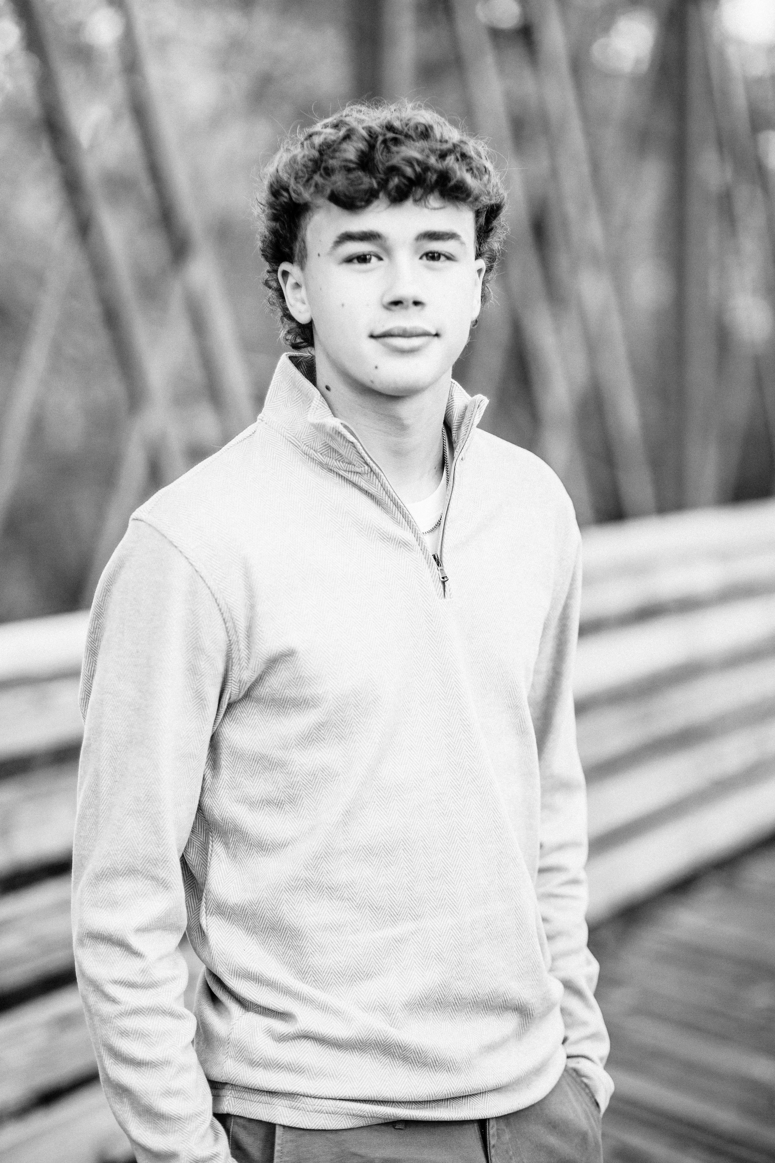 Black and white portrait of a teenage boy in a quarter-zip sweatshirt standing on a wooden bridge outdoors.