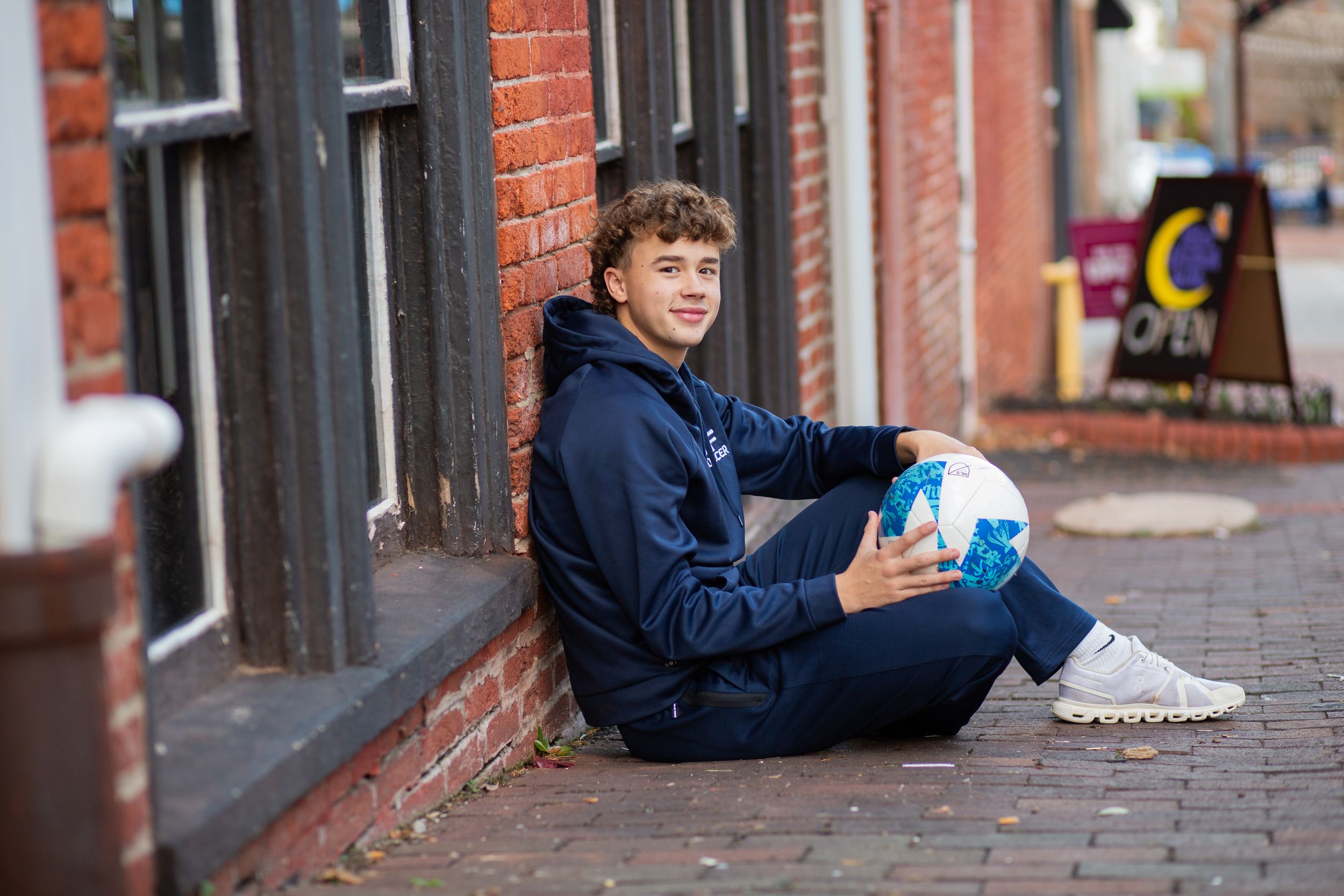 Teen boy in navy hoodie sits against brick wall holding soccer ball on cobblestone street.