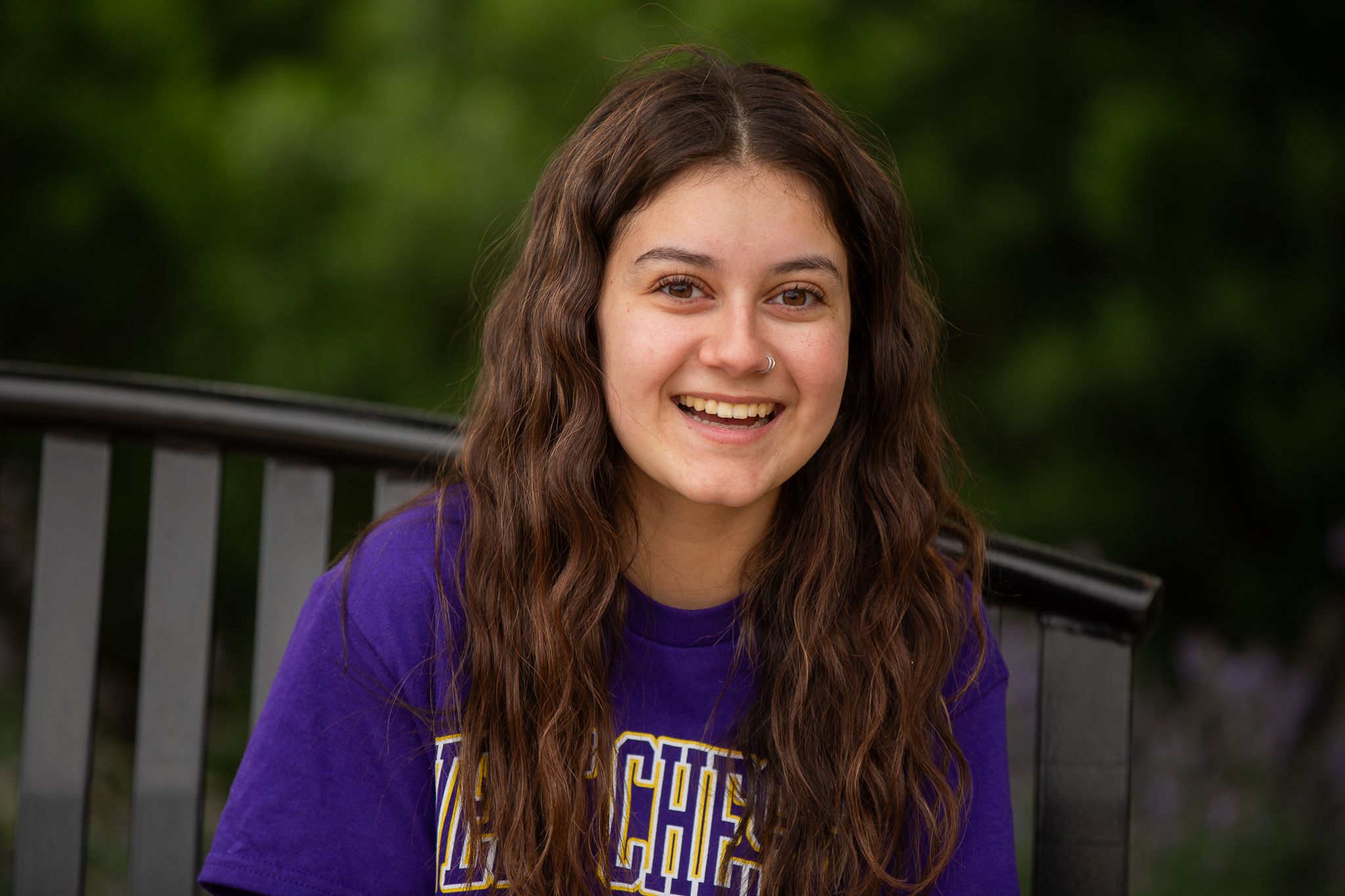 Smiling young woman with long brown hair wearing a purple college t-shirt, seated outdoors on a bench.