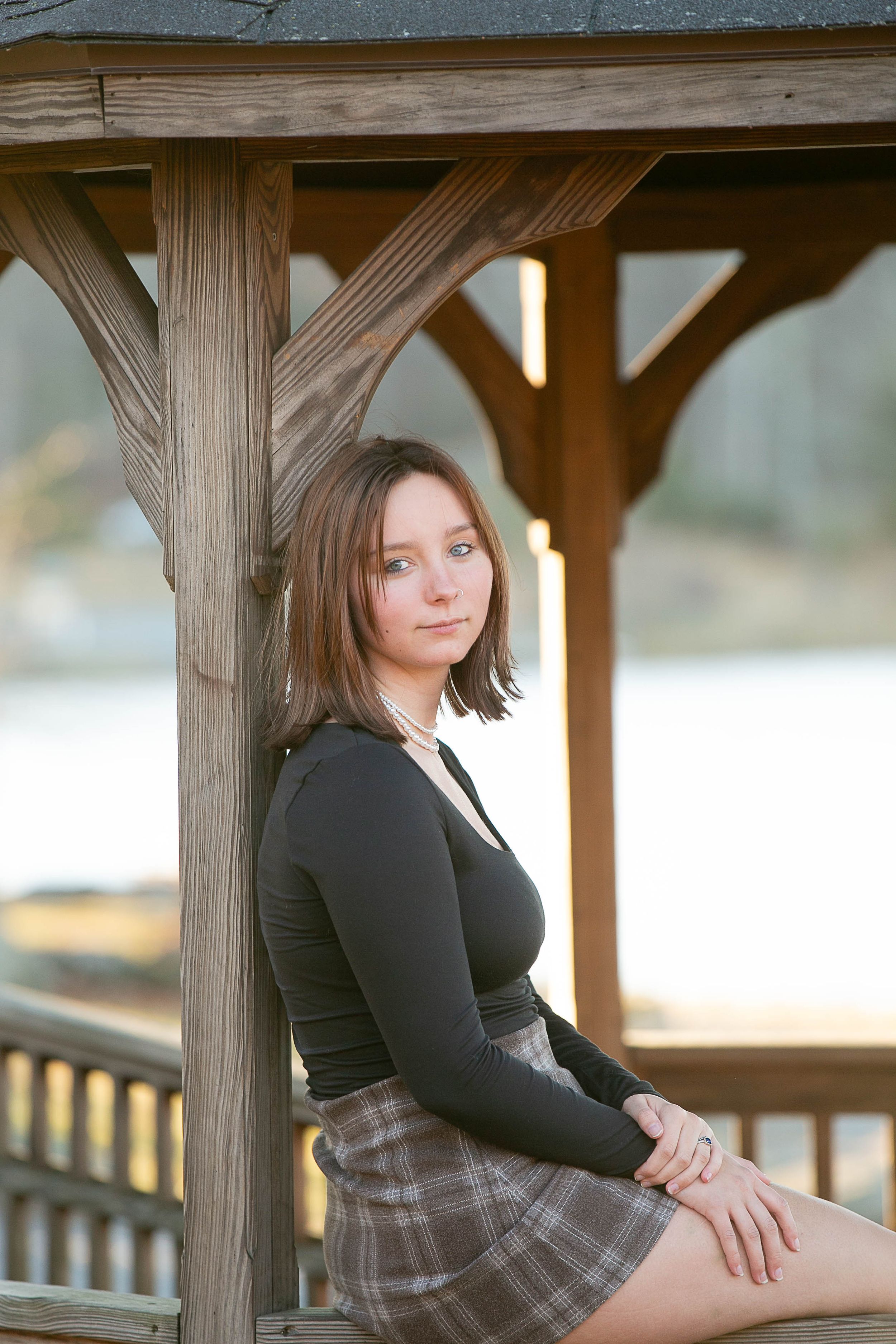 Young woman in black top and plaid skirt leaning against wooden gazebo post near a lake.