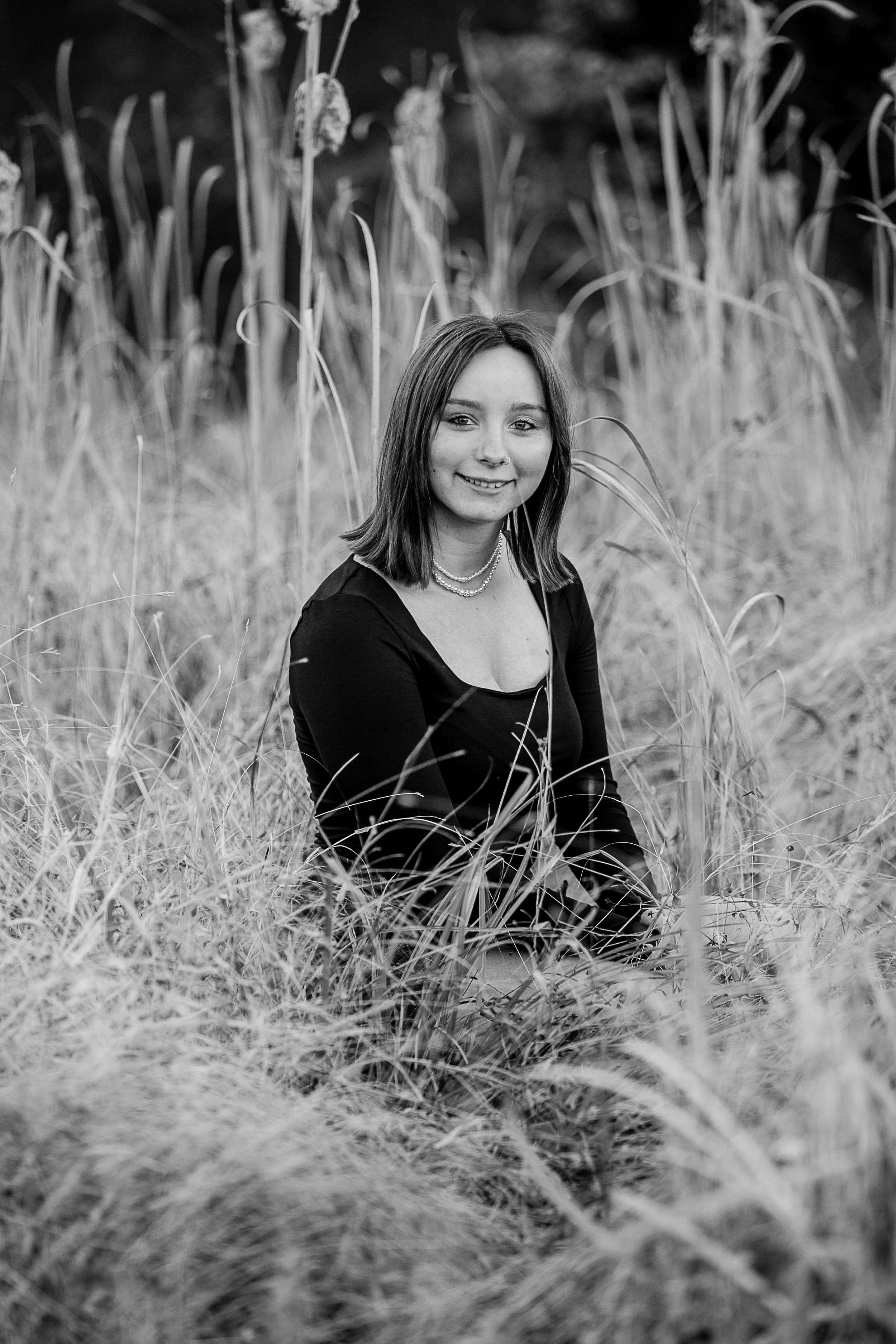 Black and white portrait of a young woman sitting in tall grass, smiling softly at the camera.