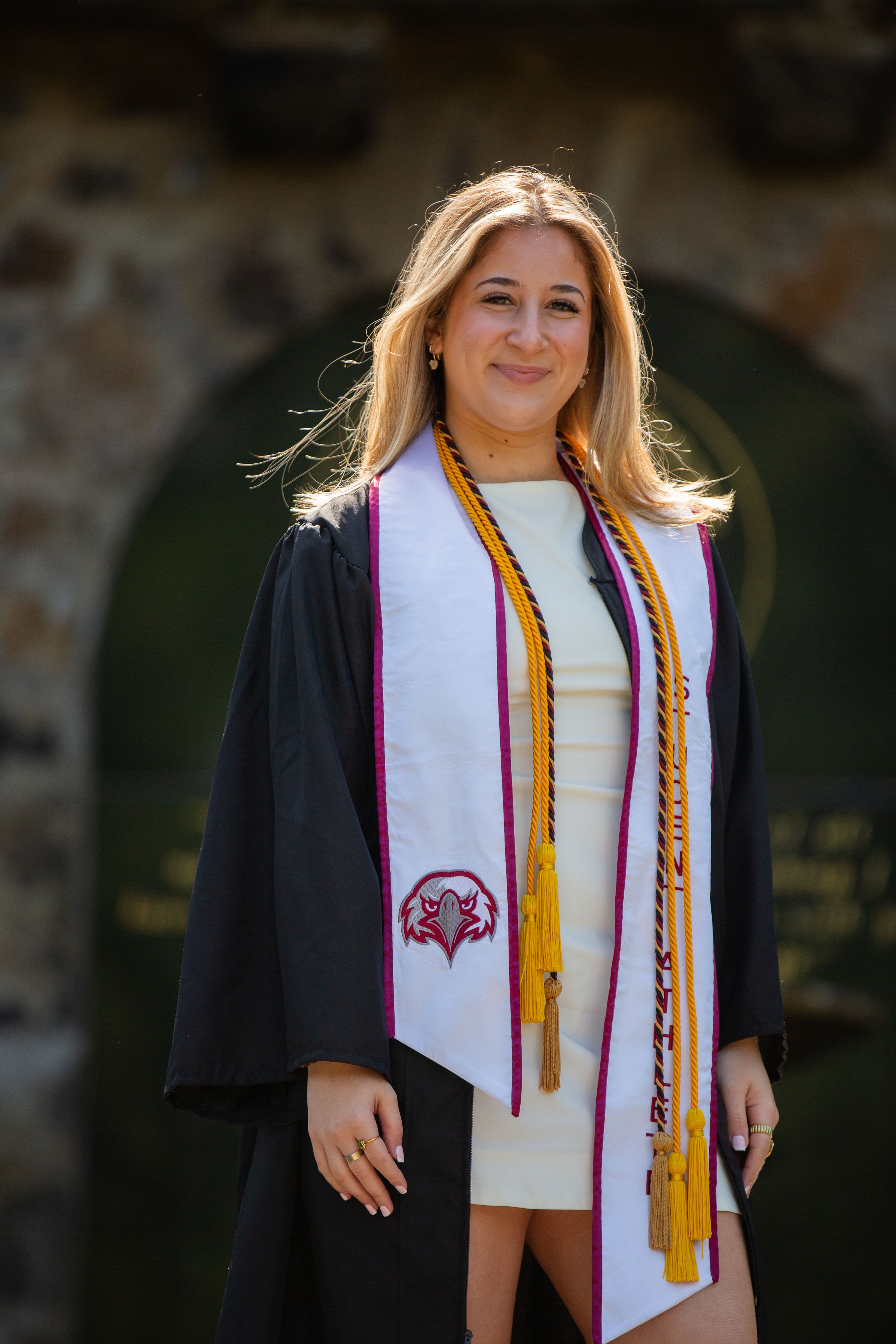 Smiling female graduate in black gown and white stole with gold tassels posing outdoors at graduation ceremony.