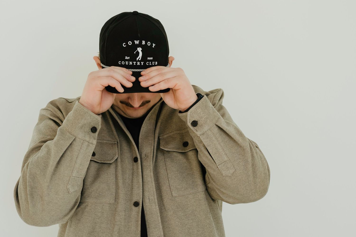 Young man with mustache wearing black snapback cap and beige jacket poses outdoors near a building.