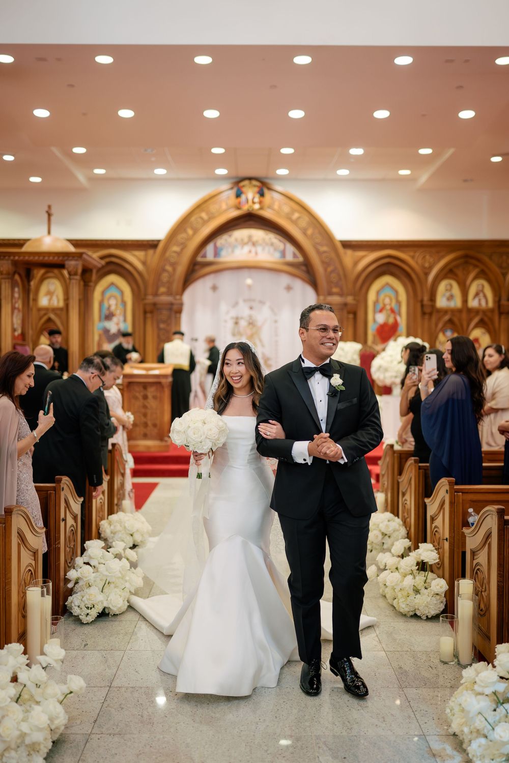 Bride and groom walk down church aisle after wedding ceremony, surrounded by white floral arrangements and guests.