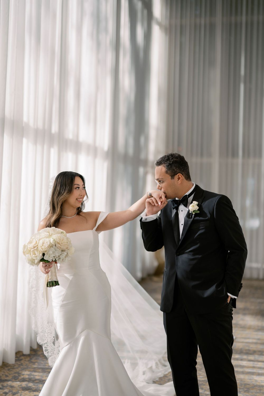 Groom in black tuxedo kisses bride's hand as she smiles holding white bouquet in elegant wedding venue.