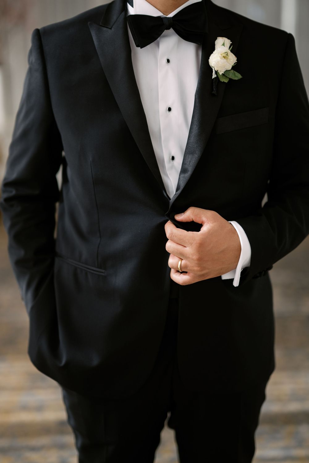 Groom in classic black tuxedo with bow tie and white rose boutonniere adjusts his jacket before the wedding.