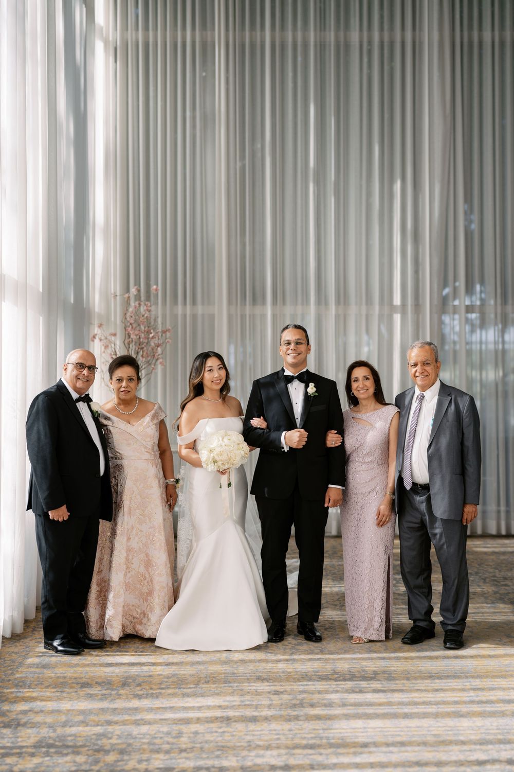 Wedding party family portrait with bride in white gown and groom in tuxedo flanked by parents.