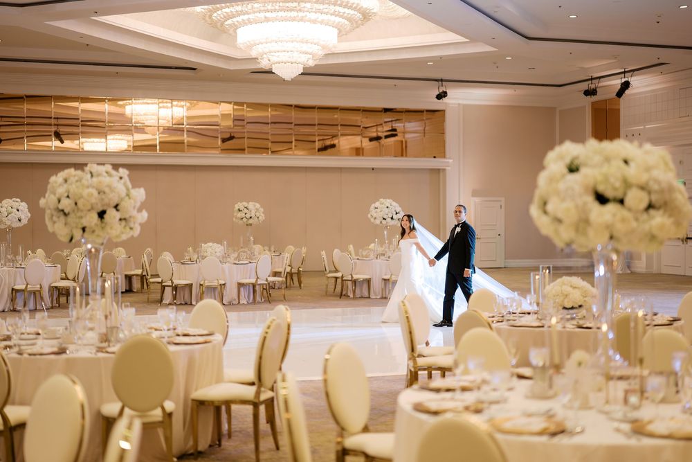 Bride and groom share first dance in elegant ballroom with tall white floral centerpieces and gold chairs at wedding reception.