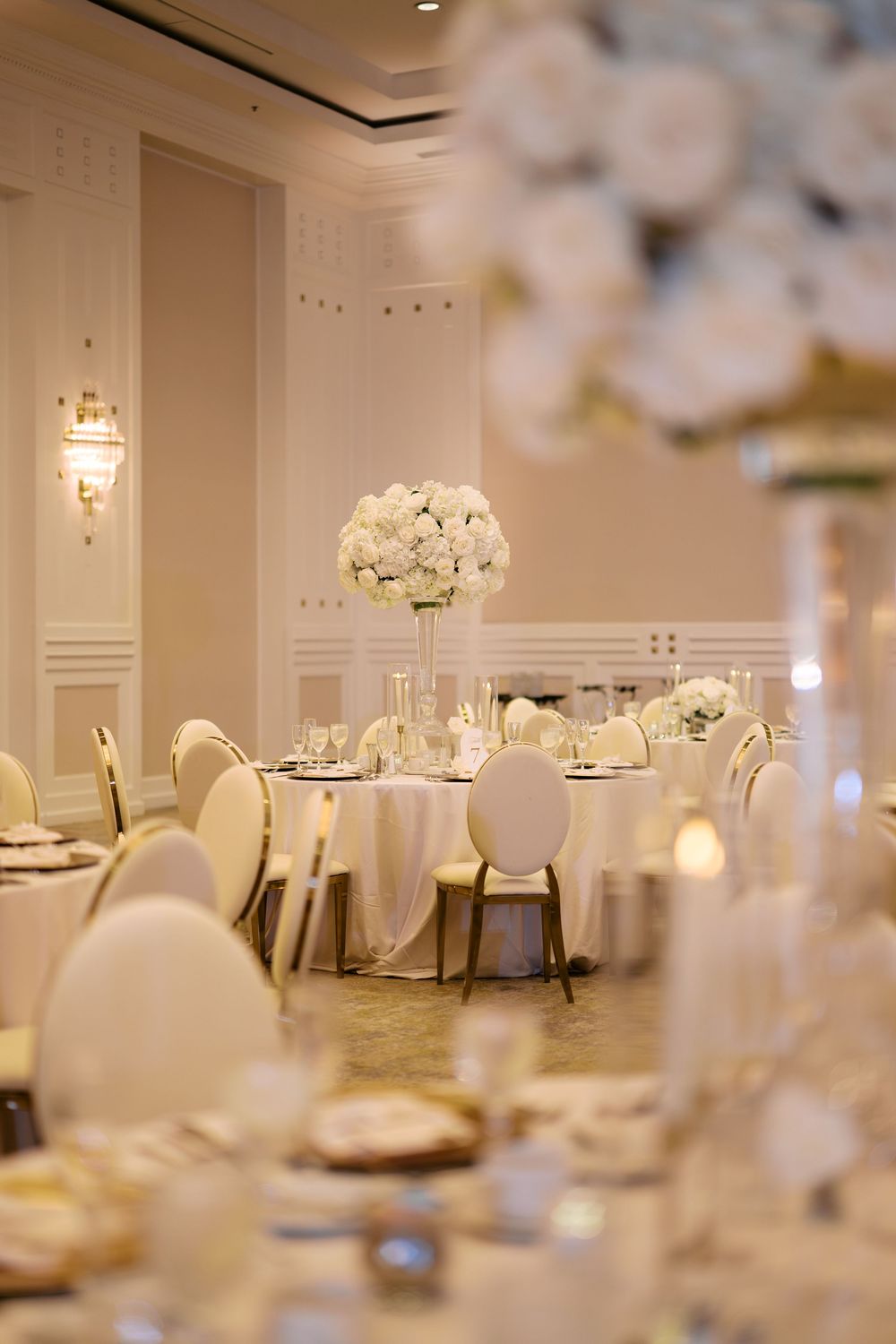 Elegant wedding reception ballroom with white floral centerpieces, round tables, gold chairs, and soft warm lighting.