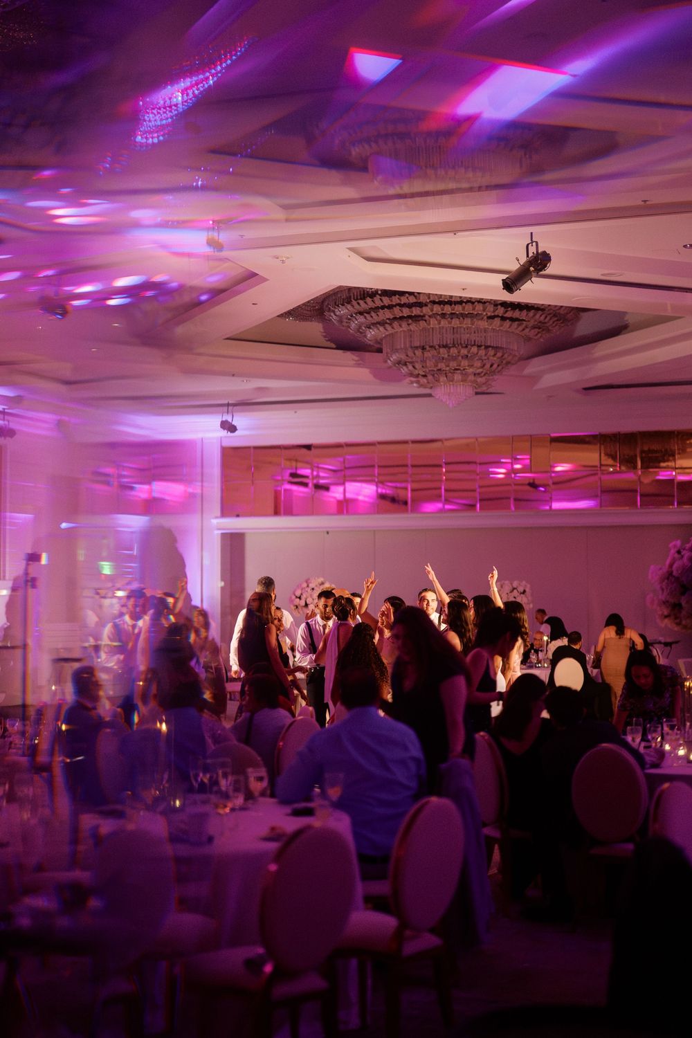 Elegant wedding reception ballroom with purple uplighting, crystal chandelier, and guests dancing on the dance floor.