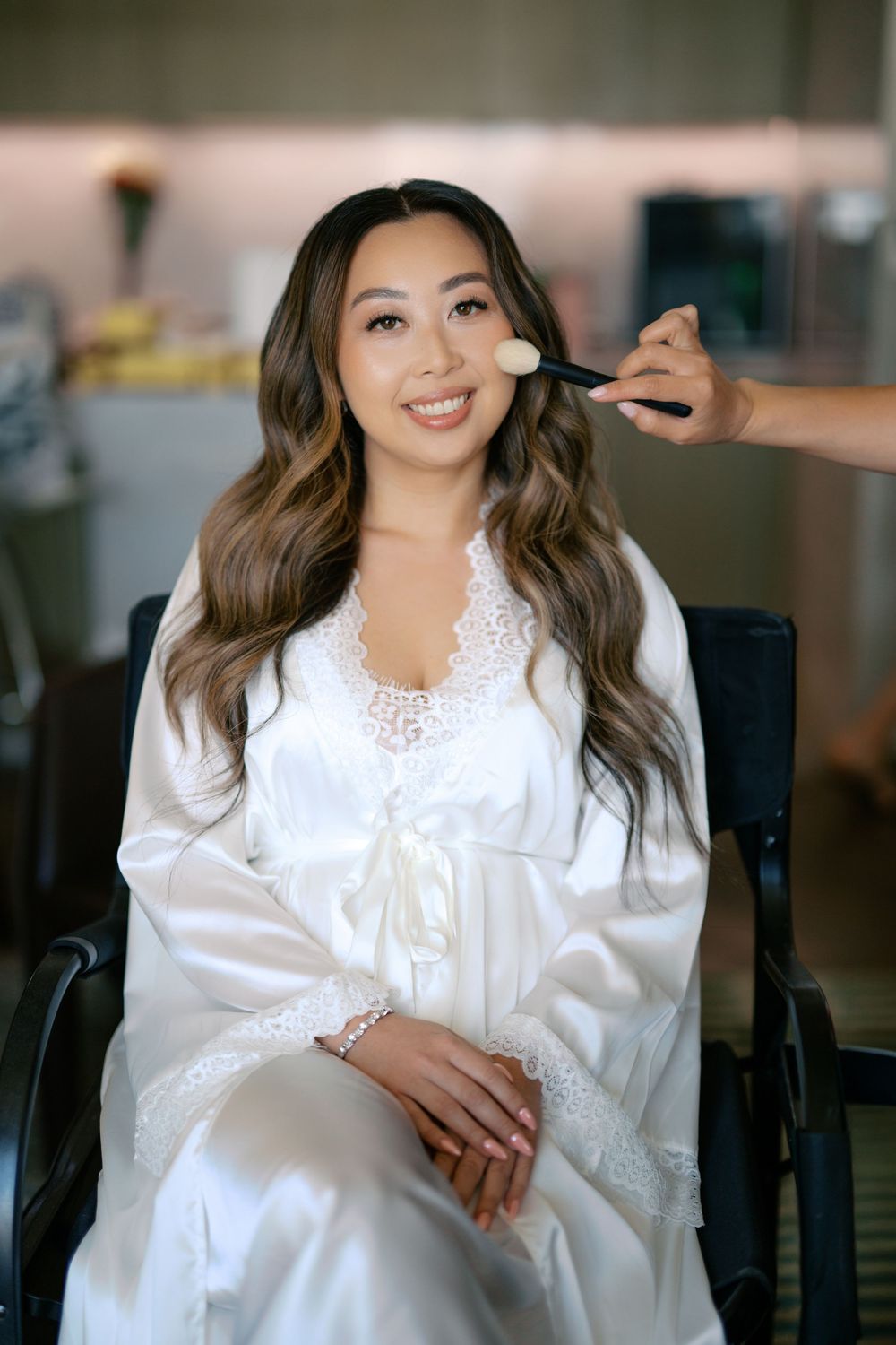 Smiling bride in white satin robe getting makeup applied before her wedding ceremony.