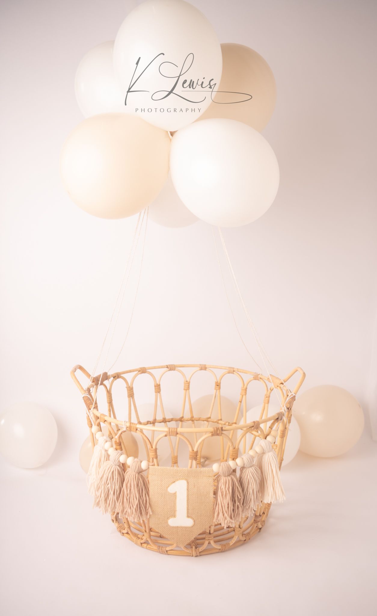 Boho-style wicker basket with number 1 and tassels, floating with white and cream balloons for a first birthday photo.