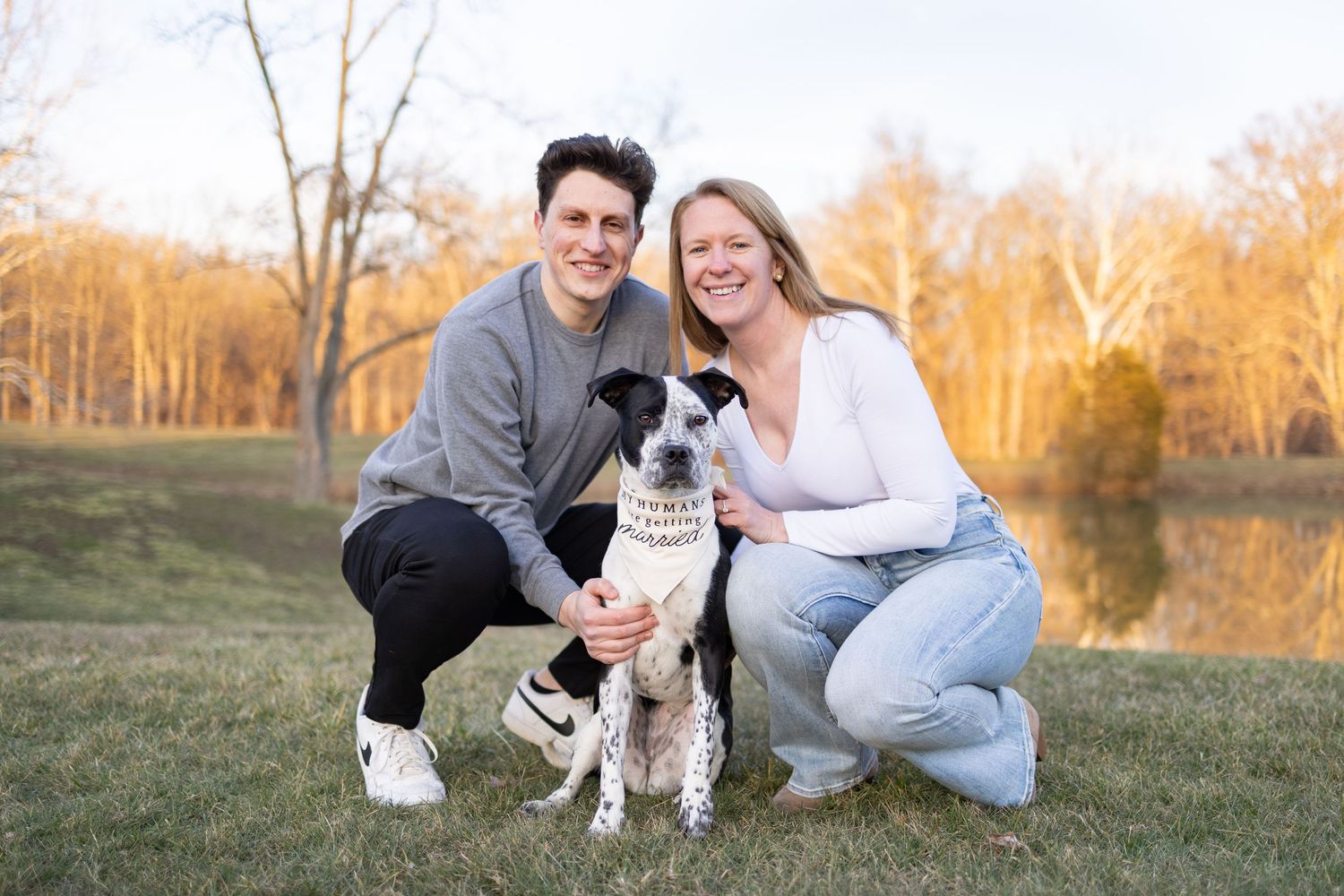 Couple crouching with spotted dog wearing bandana in autumn park near pond at golden hour.