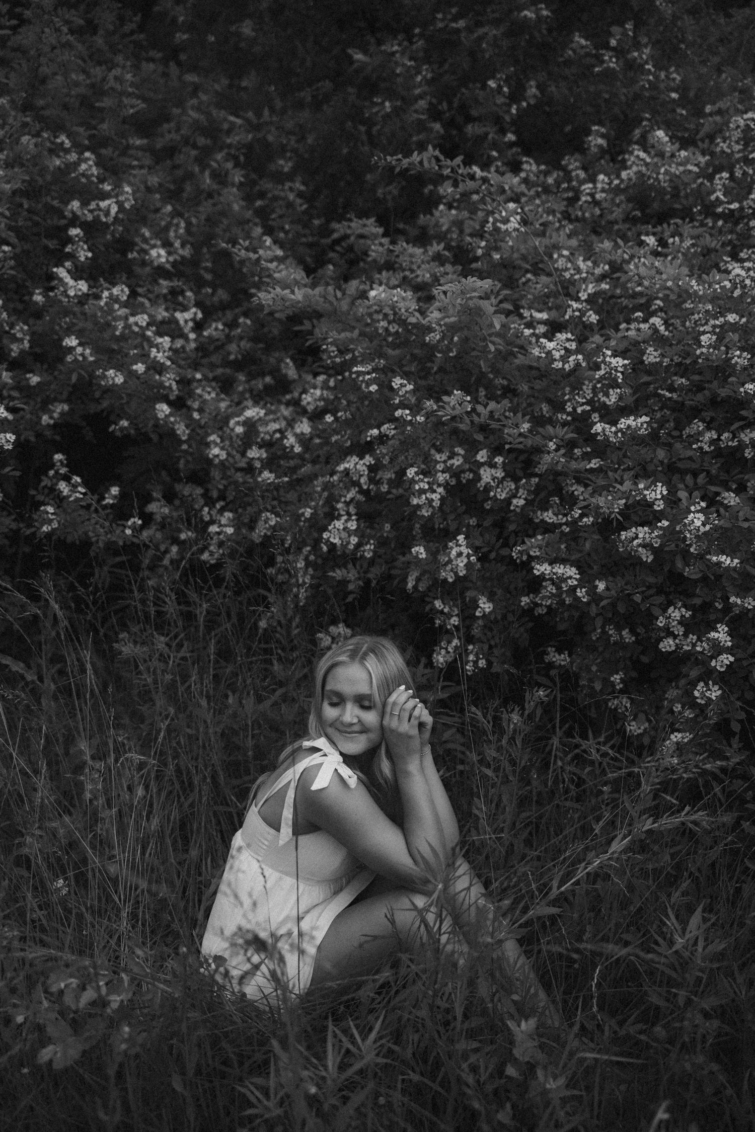 Black and white photo of a smiling blonde woman sitting in tall grass with flowering bushes behind her.