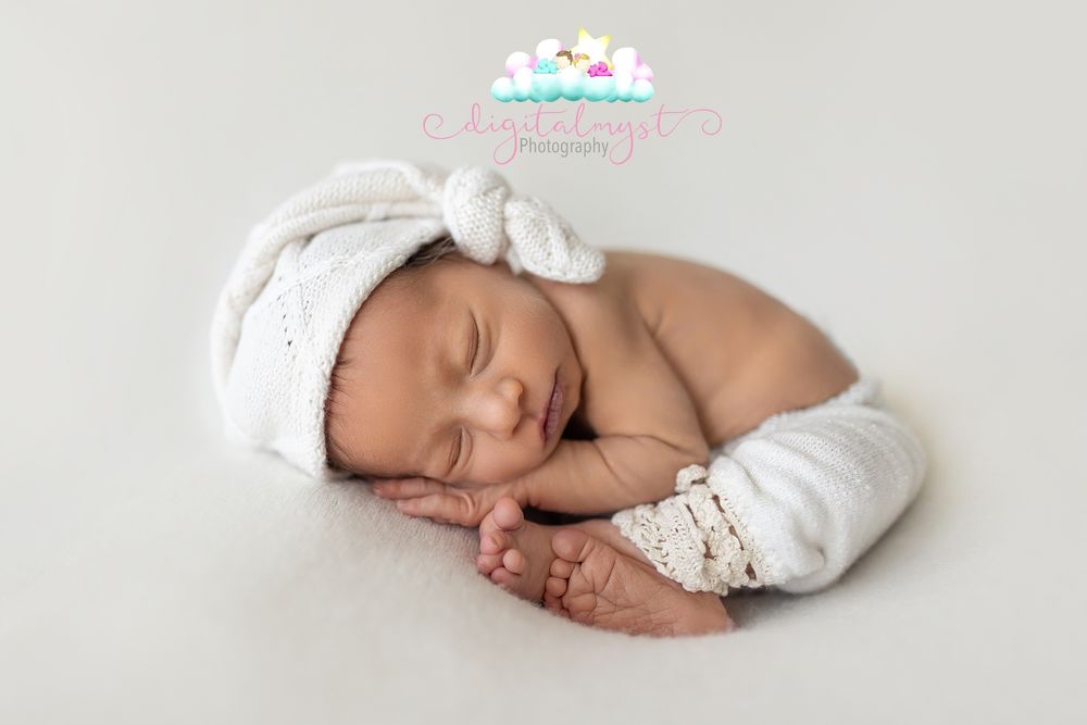 Sleeping newborn baby wearing a white knit hat and pants, curled up peacefully on a soft white background.