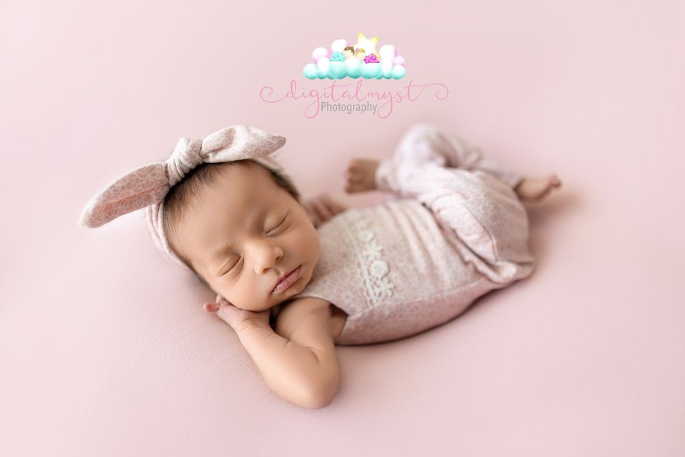 Newborn baby girl sleeping peacefully in pink romper and bunny ear headband on soft pink backdrop.
