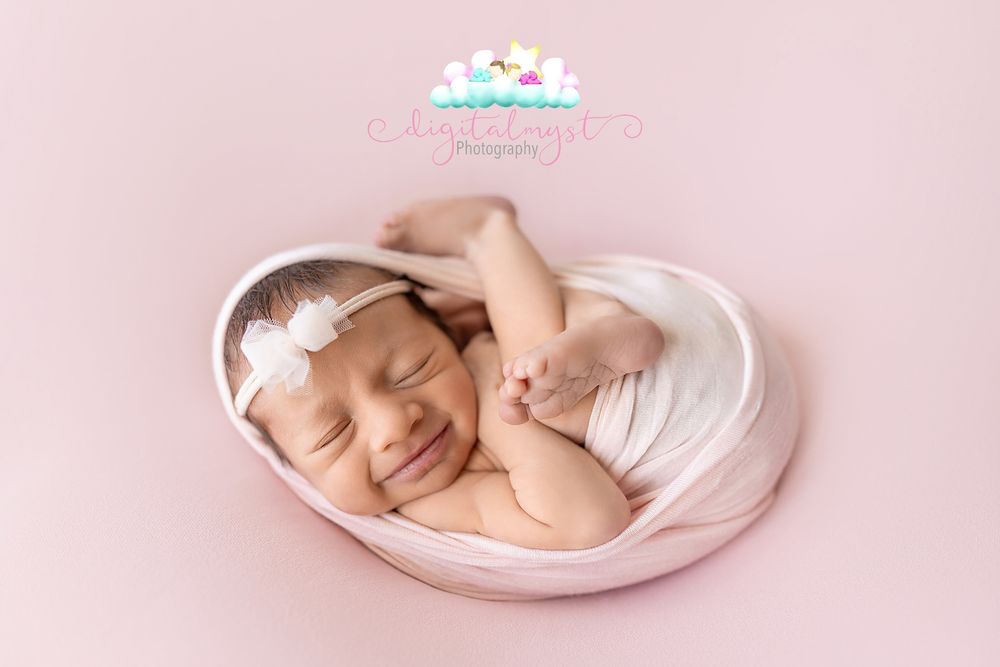 Sleeping newborn baby girl wrapped in pink fabric with white floral headband in heart-shaped bowl on pink background.