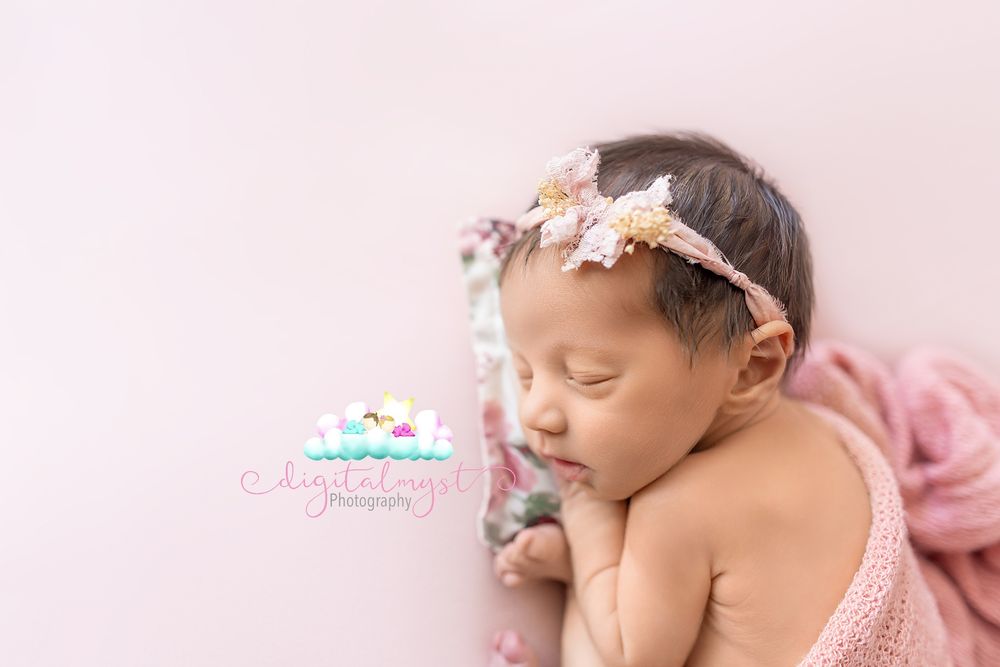 Sleeping newborn baby girl wearing a floral headband on a soft pink backdrop, professional newborn photography.