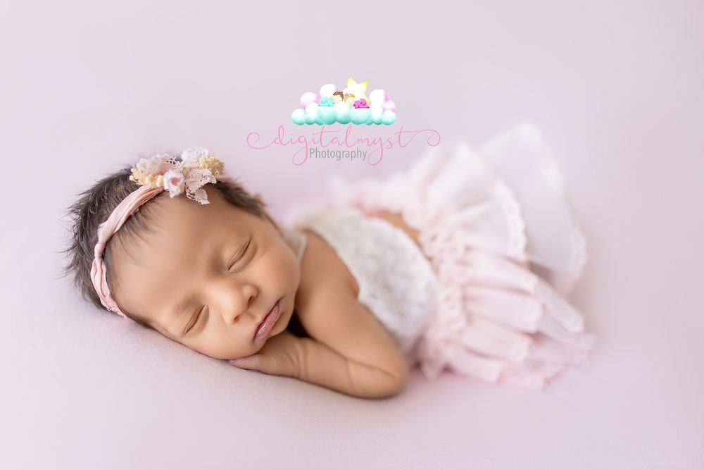 Sleeping newborn baby girl in white lace outfit and floral headband on pink background.