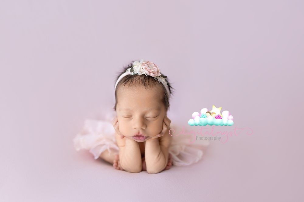 Newborn baby girl in pink tutu and floral headband posed with hands under chin on lavender backdrop.