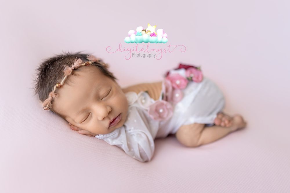 Newborn baby girl sleeping peacefully in white outfit and pink floral headband on soft pink backdrop.