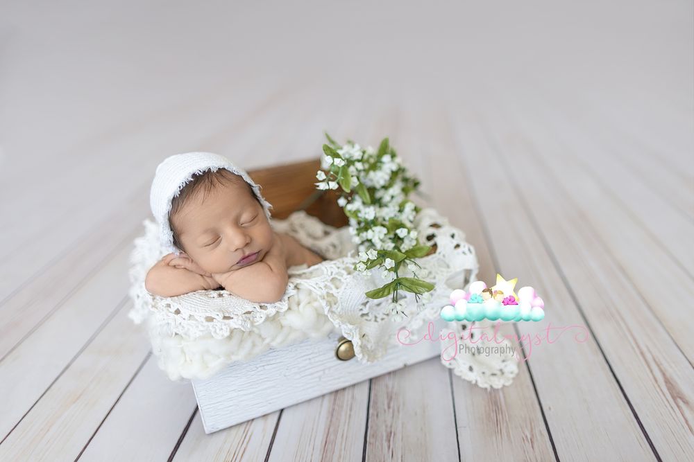 Newborn baby sleeping peacefully in white wooden box with lace and white flowers, wearing a white bonnet.