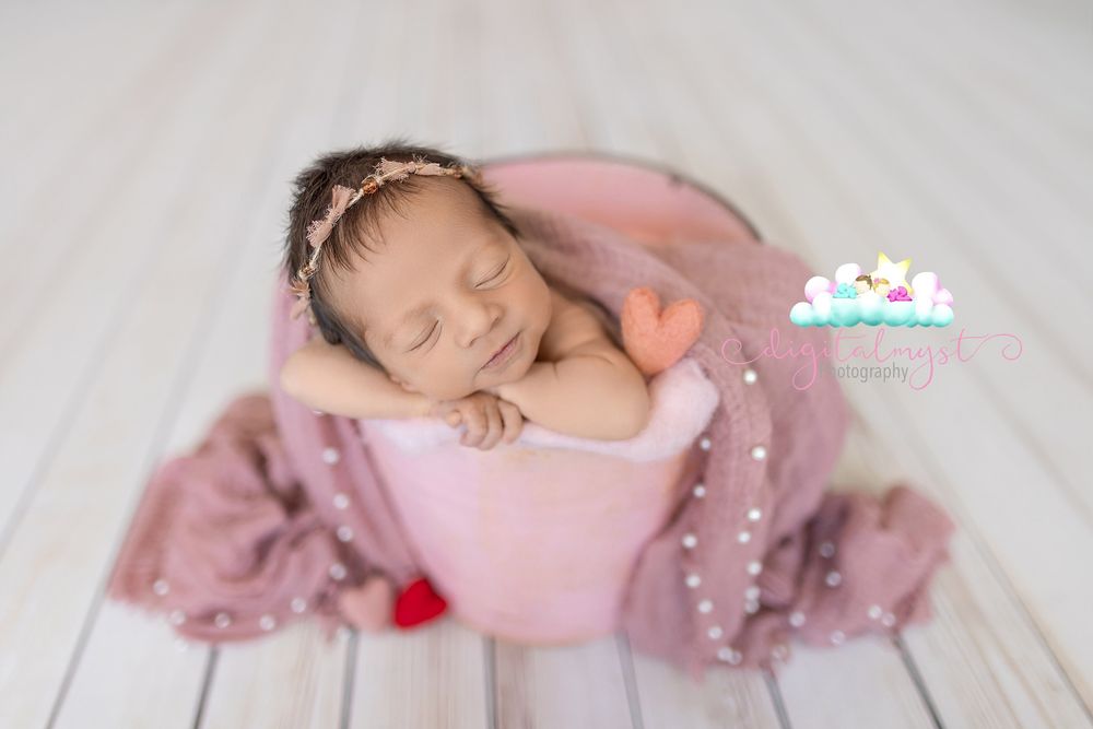 Sleeping newborn baby girl in pink wrap and floral headband posed in a blush bucket for a professional newborn photo.